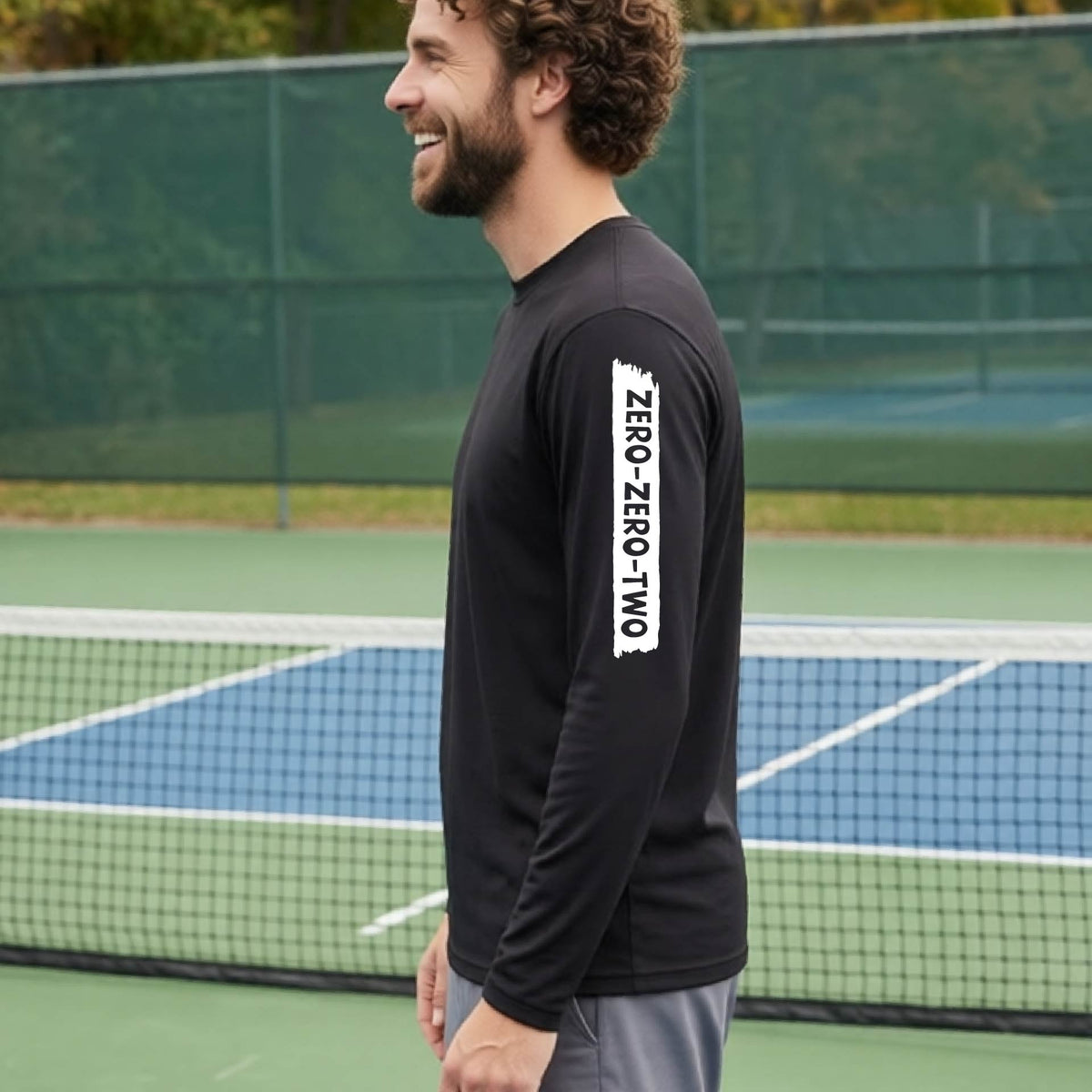 A close-up of a man on a pickleball wearing a black long-sleeve t-shirt, with a white graphic of artistic banner with the cutout words of ‘ZERO ZERO TWO’, on the sleeve of the shirt. 