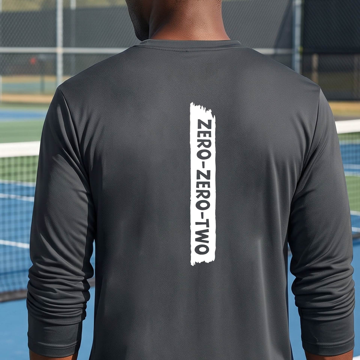 A close-up of a man on a pickleball wearing agray long-sleeve t-shirt, with a white graphic of artistic banner with the cutout words of ‘ZERO ZERO TWO’, on the back of the shirt. 