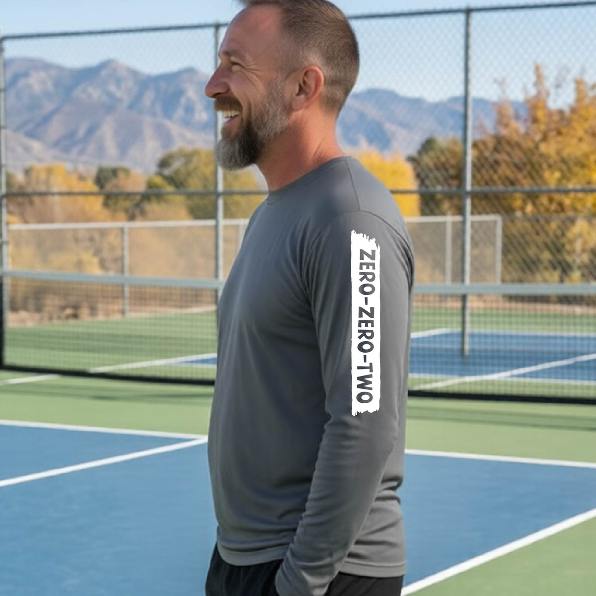A close-up of a man on a pickleball wearing agray long-sleeve t-shirt, with a white graphic of artistic banner with the cutout words of ‘ZERO ZERO TWO’, on the sleeve of the shirt. 
