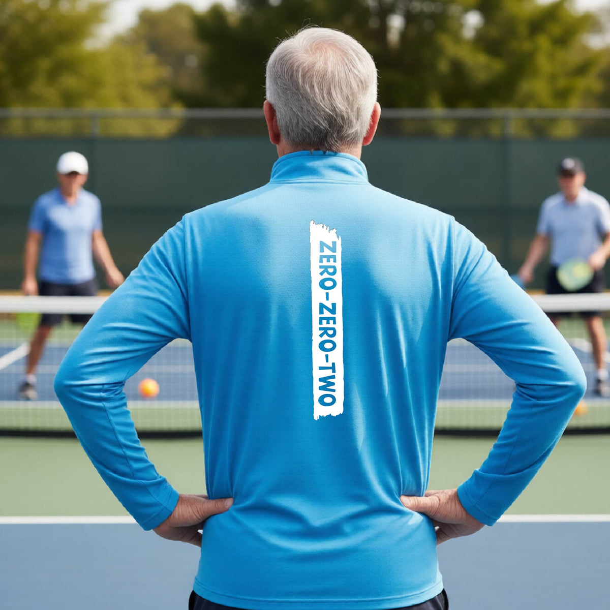 A close-up of a man, on a pickleball court wearing a brightl blue quarter-zip shirt,with a white graphic of artistic banner with the cutout words of ‘ZERO ZERO TWO’ , on the back of the shirt.