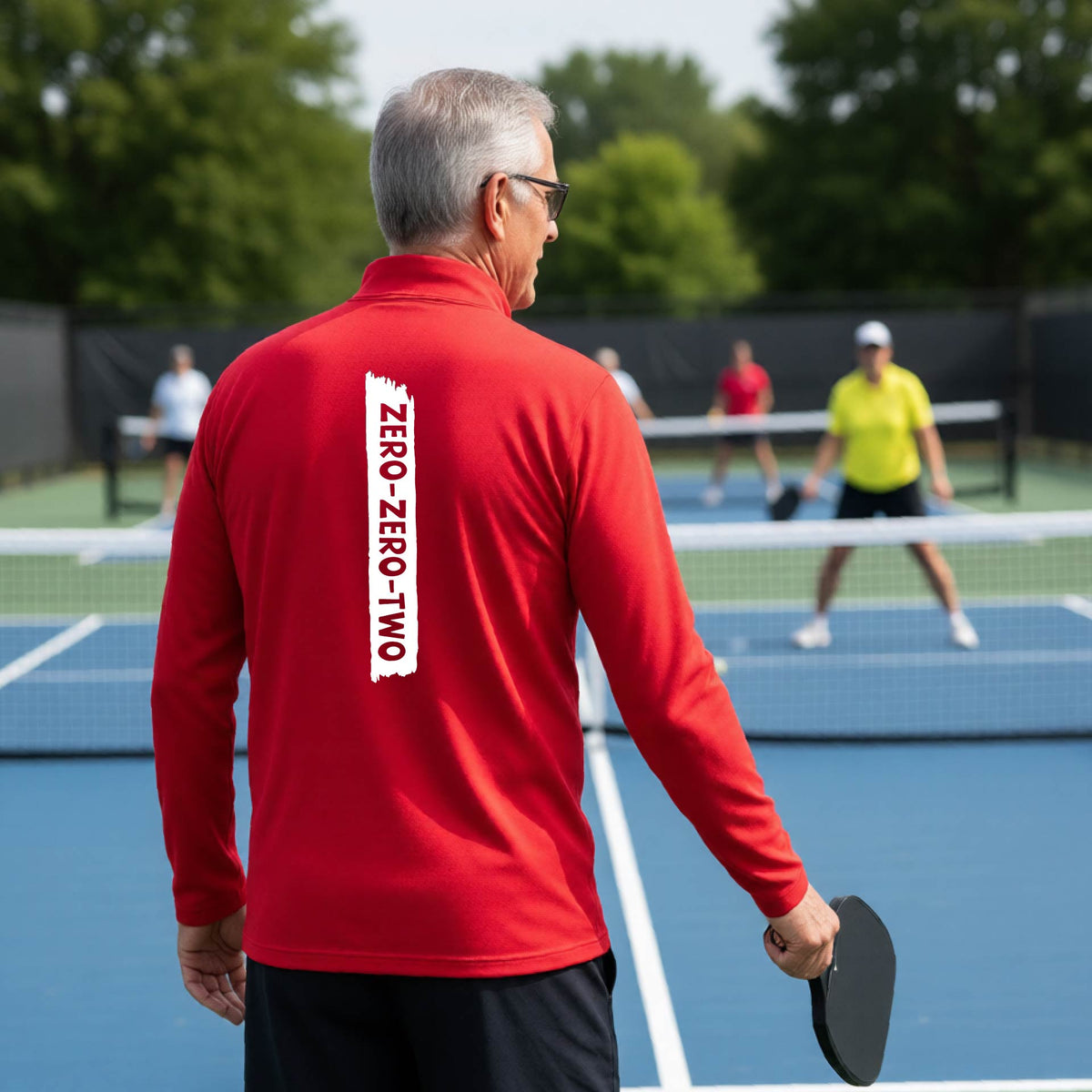 A close-up of a man, on a pickleball court wearing a red quarter-zip shirt,with a white graphic of artistic banner with the cutout words of ‘ZERO ZERO TWO’ , on the back of the shirt.