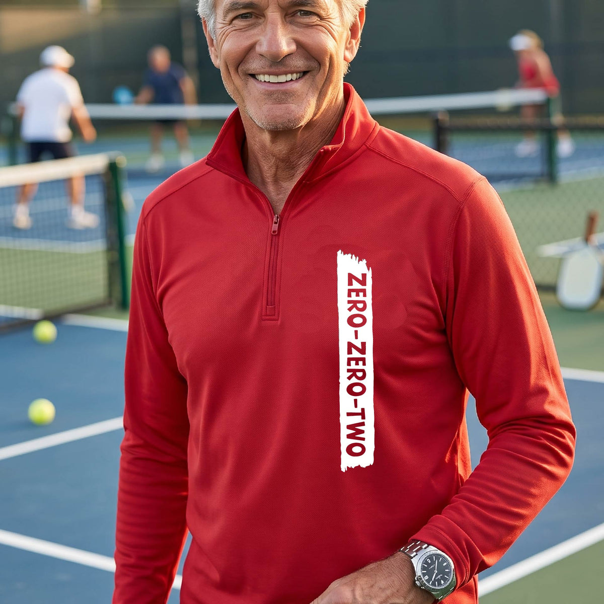 A close-up of a man, on a pickleball court wearing a red quarter-zip shirt,with a white graphic of artistic banner with the cutout words of ‘ZERO ZERO TWO’ , on the front of the shirt.