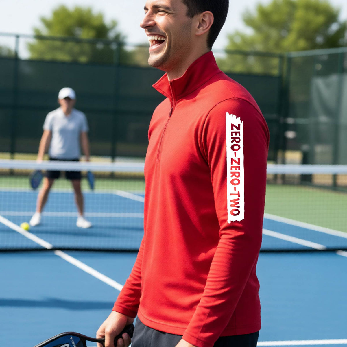 A close-up of a man, on a pickleball court wearing a red quarter-zip shirt,with a white graphic of artistic banner with the cutout words of ‘ZERO ZERO TWO’ , on the sleeve of the shirt.