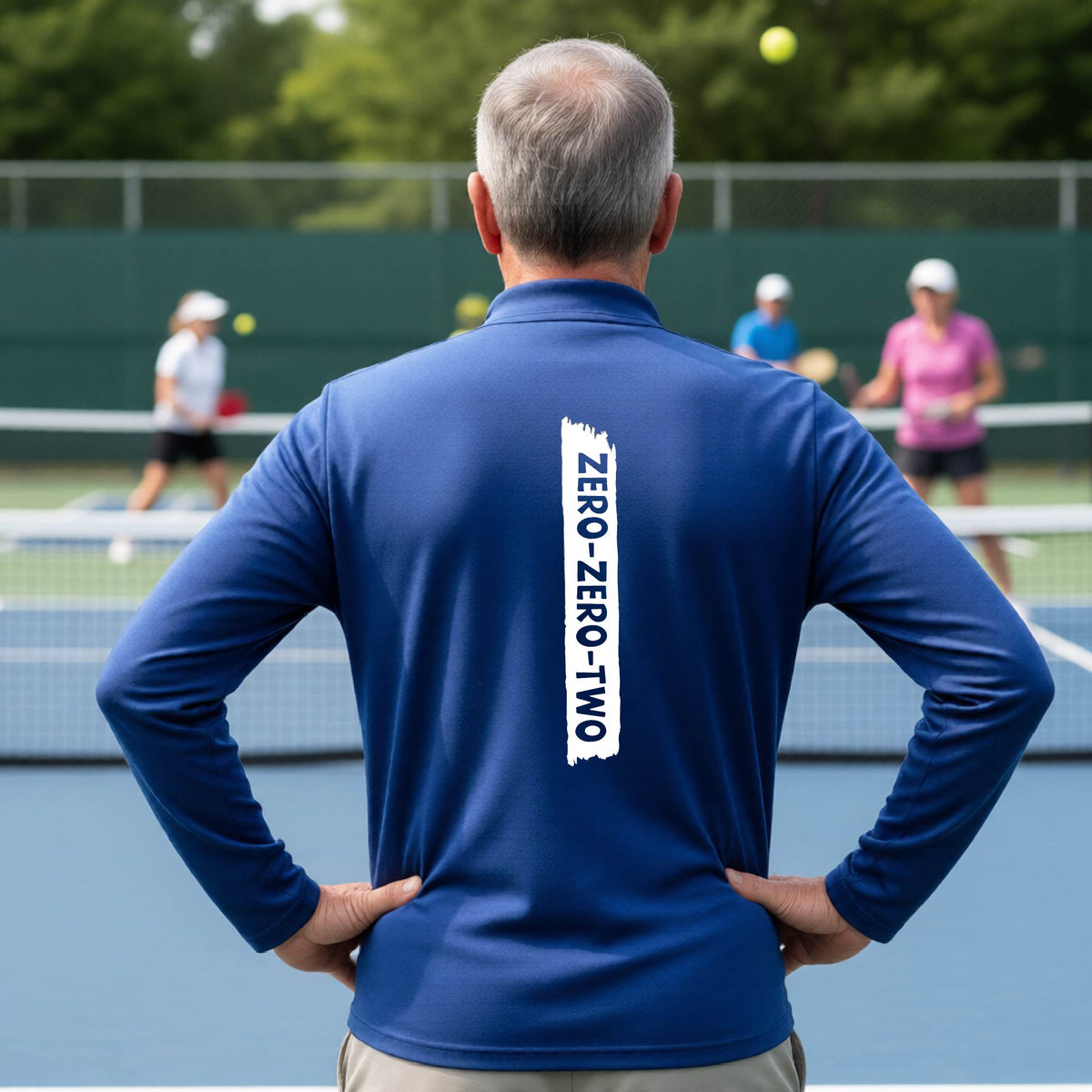 A close-up of a man, on a pickleball court wearing a royal blue quarter-zip shirt,with a white graphic of artistic banner with the cutout words of ‘ZERO ZERO TWO’ , on the back of the shirt.