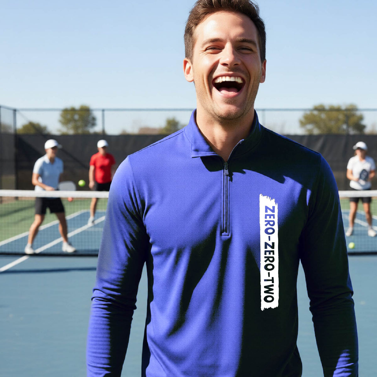 A close-up of a man, on a pickleball court wearing a royal blue quarter-zip shirt,with a white graphic of artistic banner with the cutout words of ‘ZERO ZERO TWO’ , on the front of the shirt.