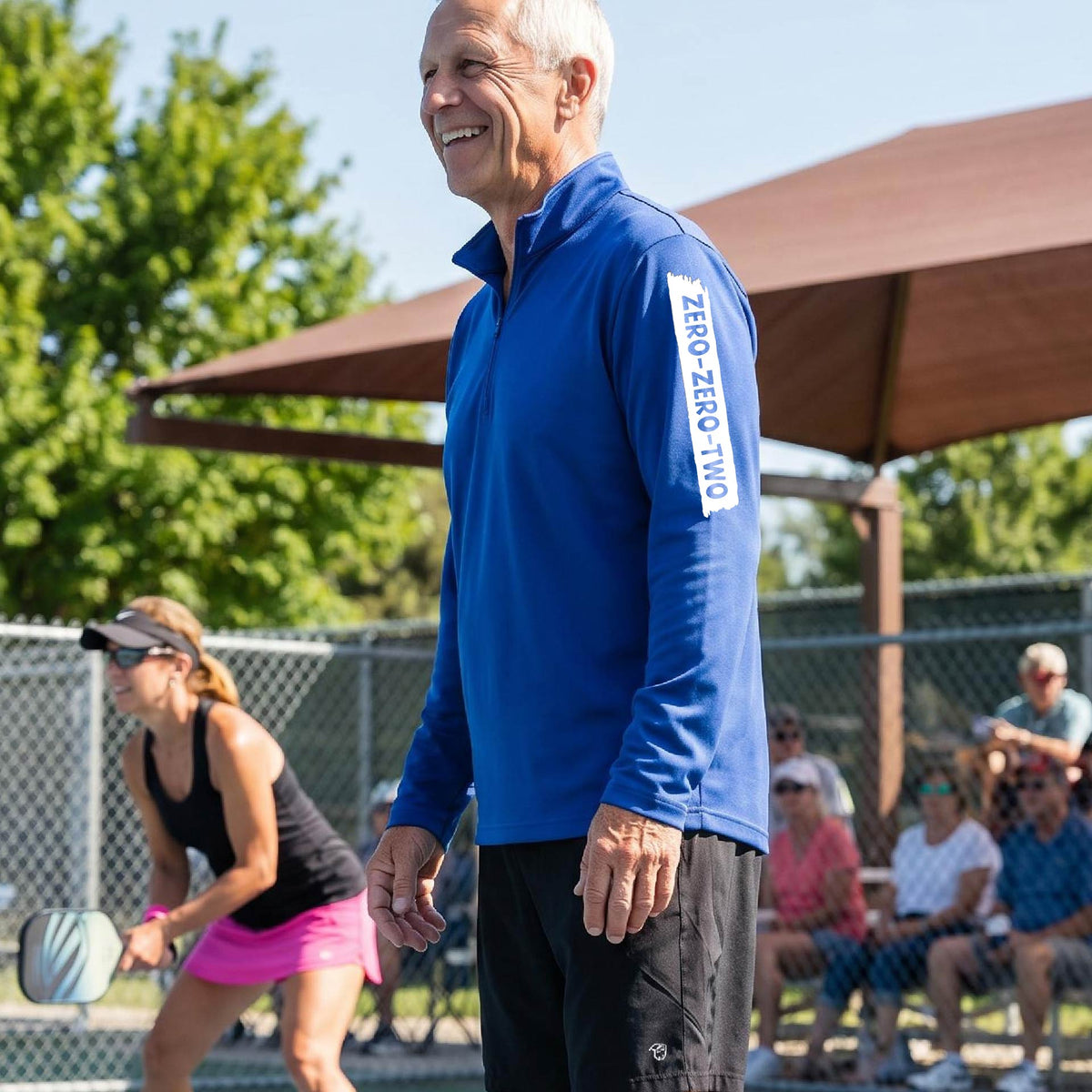 A close-up of a man, on a pickleball court wearing a royal blue quarter-zip shirt,with a white graphic of artistic banner with the cutout words of ‘ZERO ZERO TWO’ , on the sleeve of the shirt.