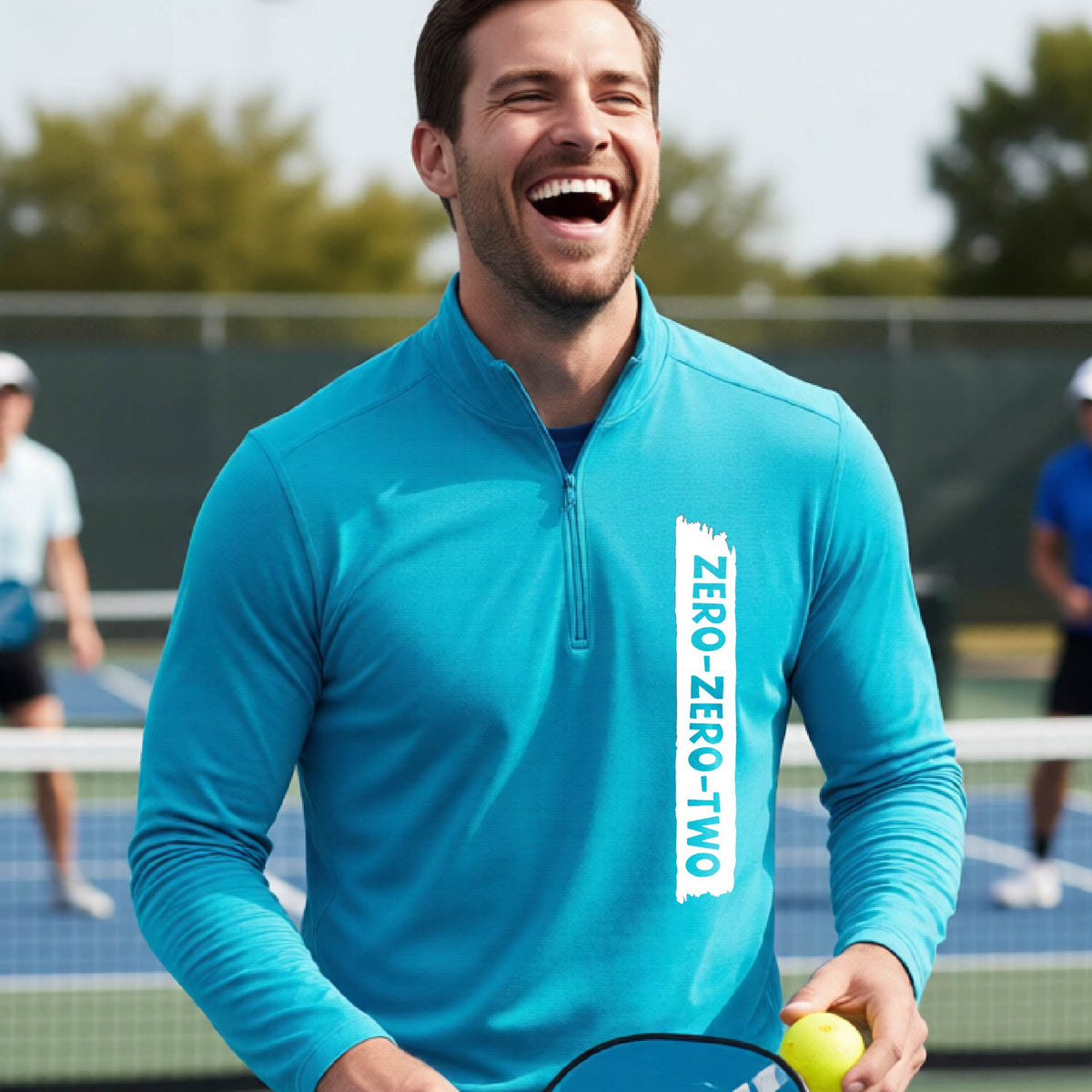 A close-up of a man, on a pickleball court wearing a brightl blue quarter-zip shirt,with a white graphic of artistic banner with the cutout words of ‘ZERO ZERO TWO’ , on the front of the shirt.