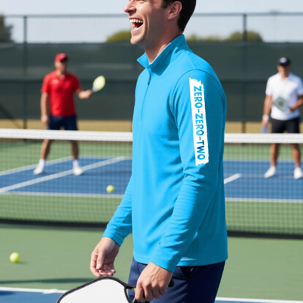 A close-up of a man, on a pickleball court wearing a brightl blue quarter-zip shirt,with a white graphic of artistic banner with the cutout words of ‘ZERO ZERO TWO’ , on the sleeve of the shirt.