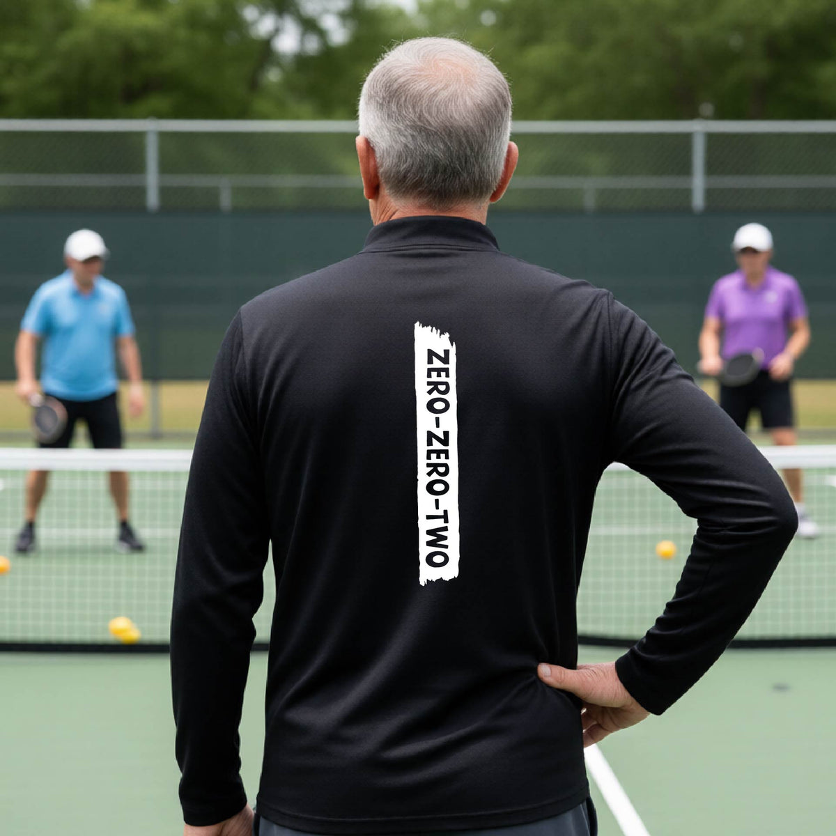 A close-up of a man, on a pickleball court wearing a black quarter-zip shirt,with a white graphic of artistic banner with the cutout words of ‘ZERO ZERO TWO’ , on the back of the shirt.