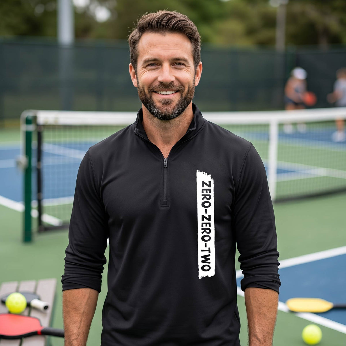 A close-up of a man, on a pickleball court wearing a black quarter-zip shirt,with a white graphic of artistic banner with the cutout words of ‘ZERO ZERO TWO’ , on the front of the shirt.