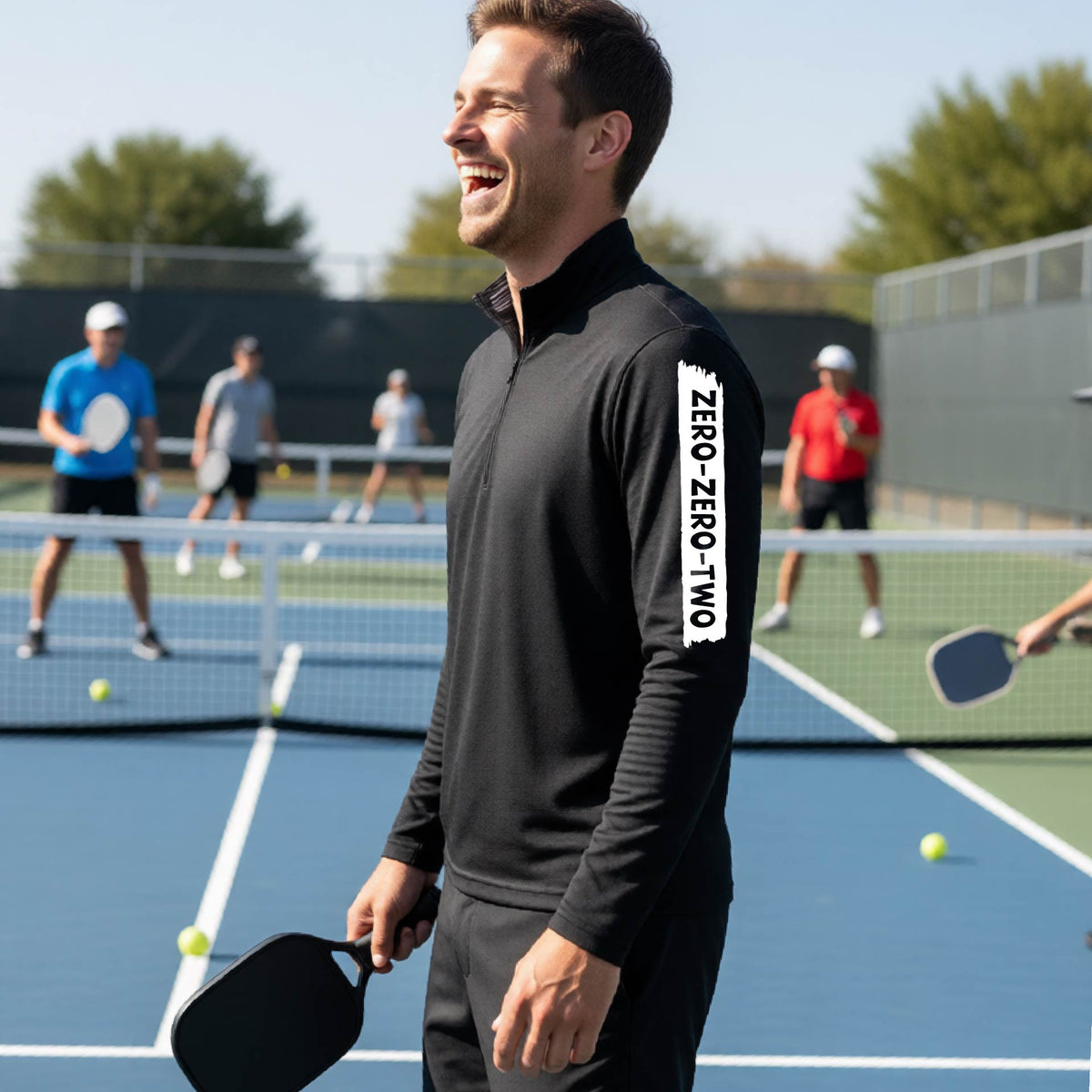 A close-up of a man, on a pickleball court wearing a black quarter-zip shirt,with a white graphic of artistic banner with the cutout words of ‘ZERO ZERO TWO’ , on the sleeve of the shirt.