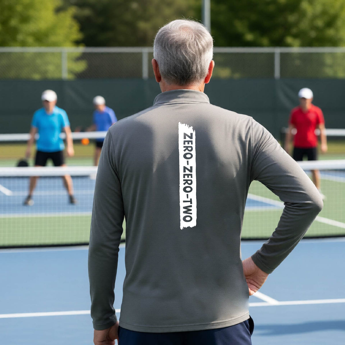A close-up of a man, on a pickleball court wearing a gray quarter-zip shirt,with a white graphic of artistic banner with the cutout words of ‘ZERO ZERO TWO’ , on the back of the shirt. 