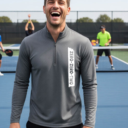 A close-up of a man, on a pickleball court wearing a gray quarter-zip shirt,with a white graphic of artistic banner with the cutout words of ‘ZERO ZERO TWO’ , on the front of the shirt.