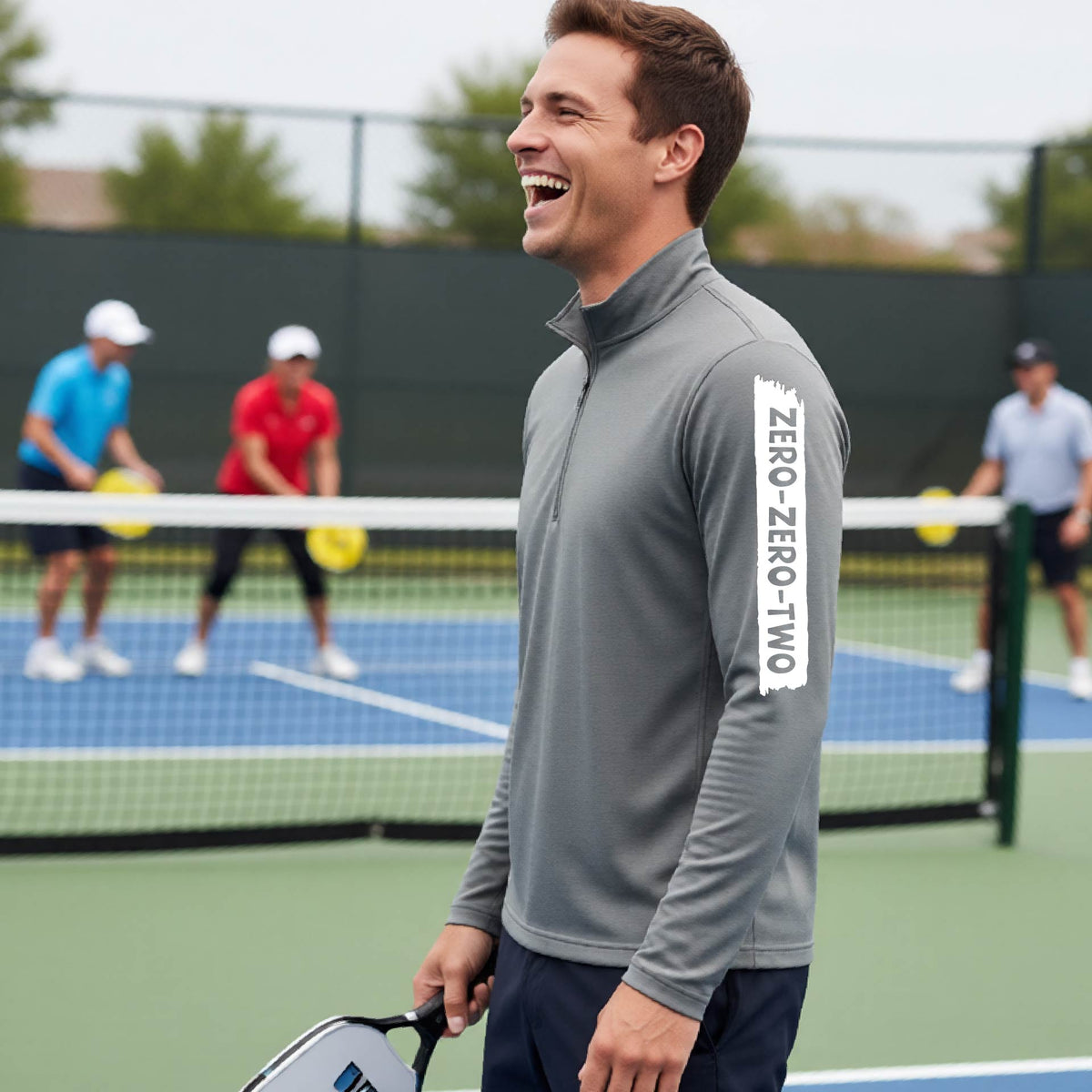 A close-up of a man, on a pickleball court wearing a gray quarter-zip shirt,with a white graphic of artistic banner with the cutout words of ‘ZERO ZERO TWO’ , on the sleeve of the shirt. 