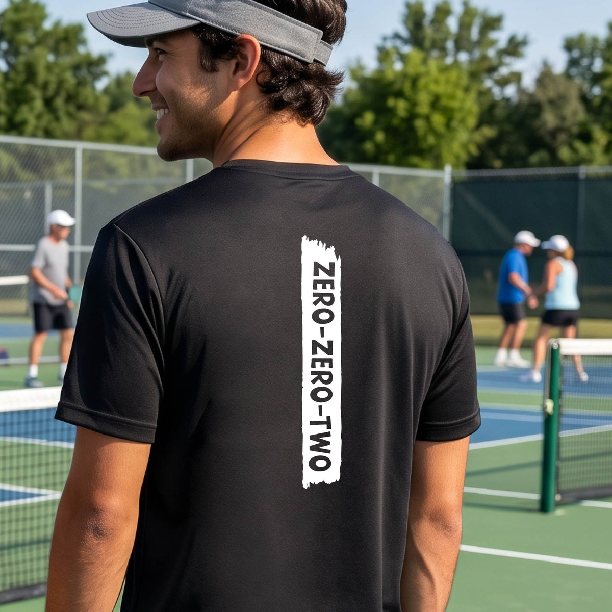A close-up of a man on a pickleball wearing a black short-sleeve t-shirt, with a white graphic of artistic banner with the cutout words of ‘ZERO ZERO TWO’, on the back of the shirt. 