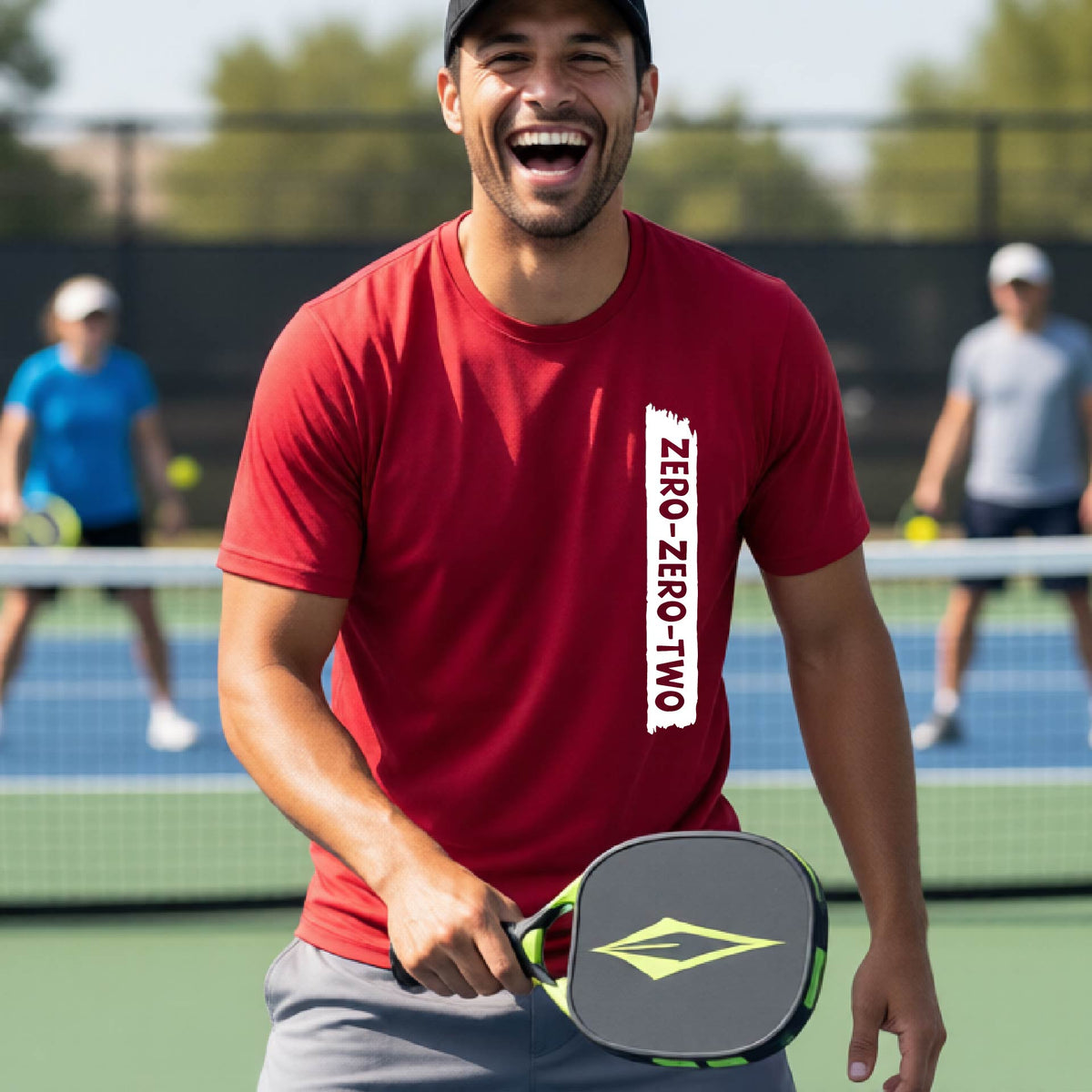 A close-up of a man on a pickleball wearing a red short-sleeve t-shirt, with a white graphic of artistic banner with the cutout words of ‘ZERO ZERO TWO’, on the front of the shirt. 