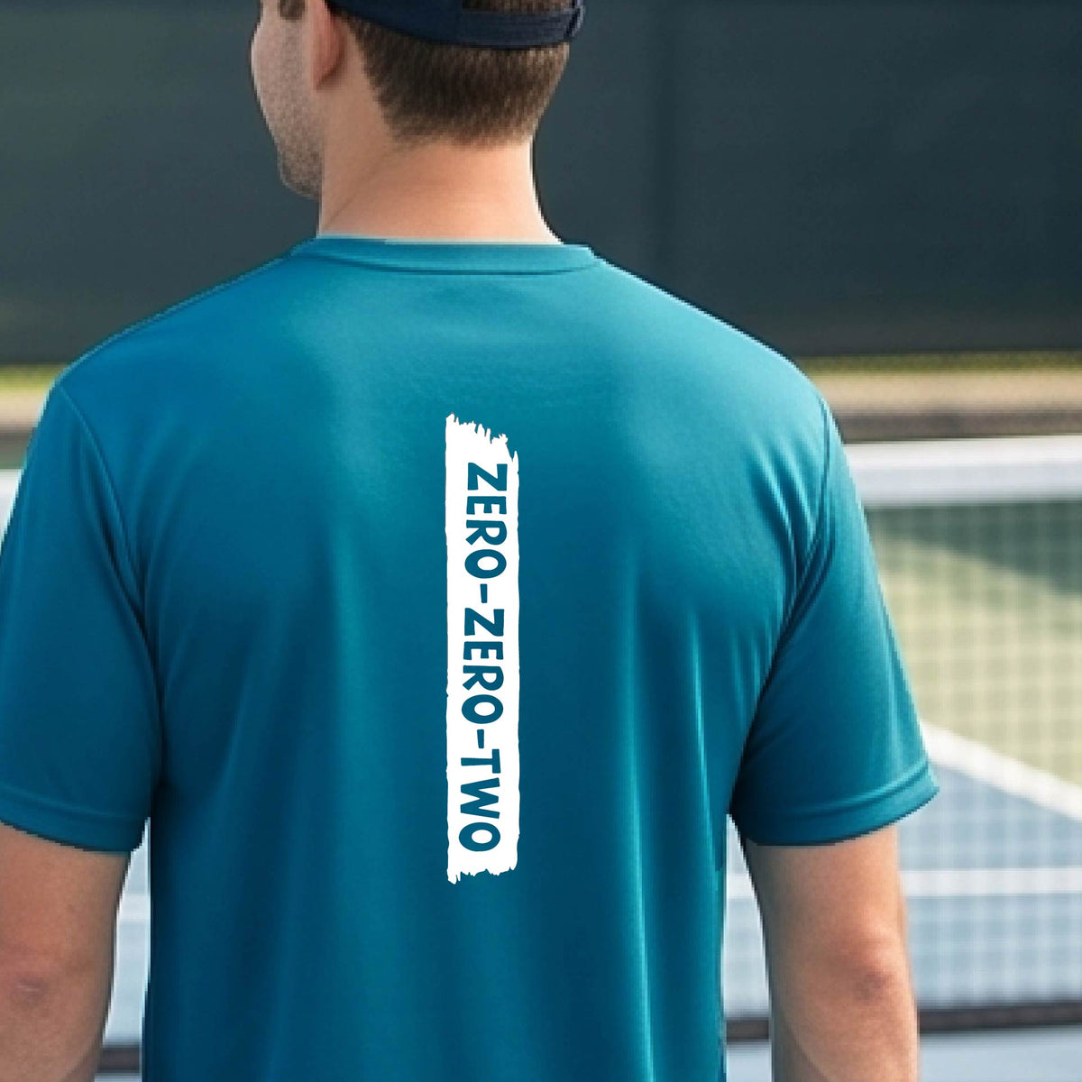 A close-up of a man on a pickleball wearing a teal short-sleeve t-shirt, with a white graphic of artistic banner with the cutout words of ‘ZERO ZERO TWO’, on the back of the shirt.
