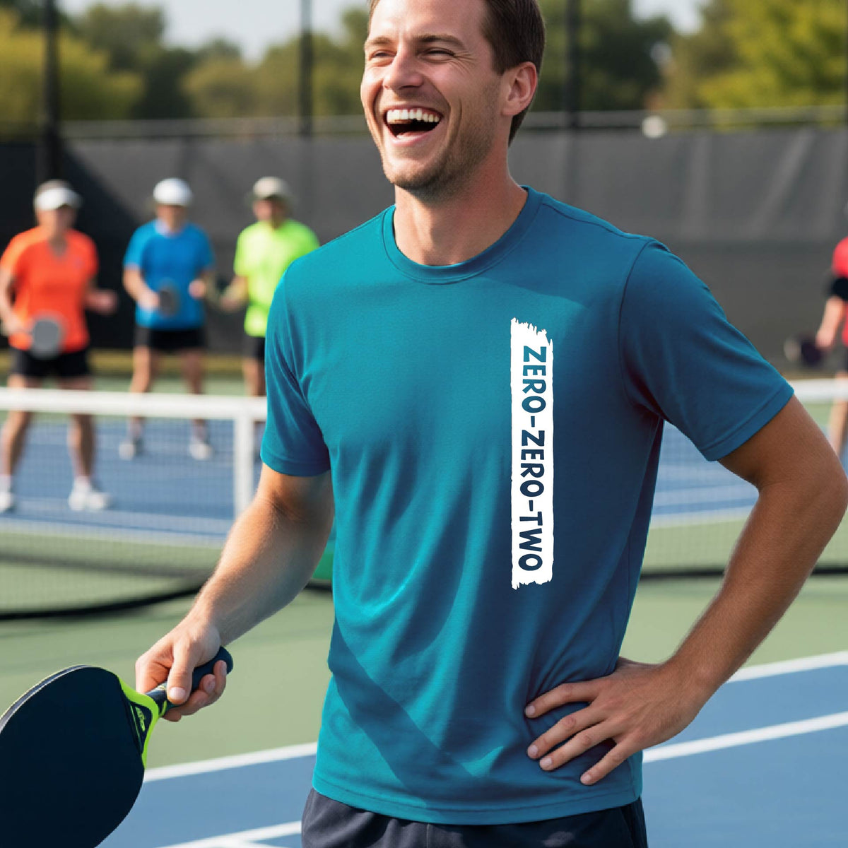 A close-up of a man on a pickleball wearing a teal short-sleeve t-shirt, with a white graphic of artistic banner with the cutout words of ‘ZERO ZERO TWO’, on the front of the shirt. 