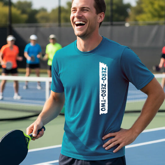 A close-up of a man on a pickleball wearing a teal short-sleeve t-shirt, with a white graphic of artistic banner with the cutout words of ‘ZERO ZERO TWO’, on the front of the shirt. 