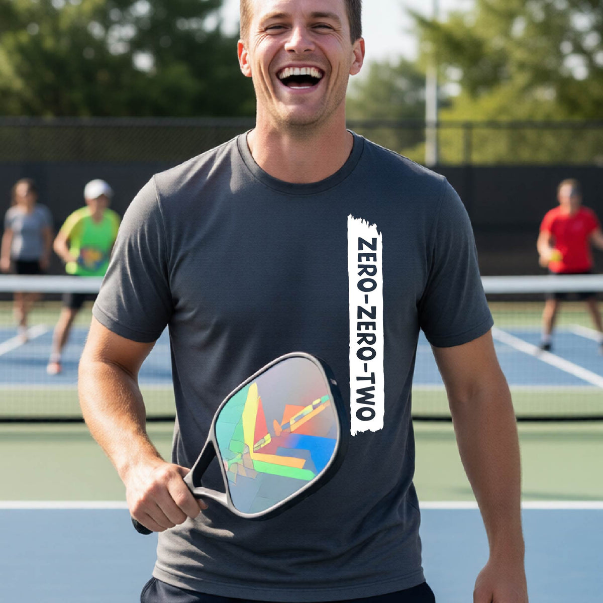 A close-up of a man on a pickleball wearing a dark gray short-sleeve t-shirt, with a white graphic of artistic banner with the cutout words of ‘ZERO ZERO TWO’, on the front of the shirt. 
