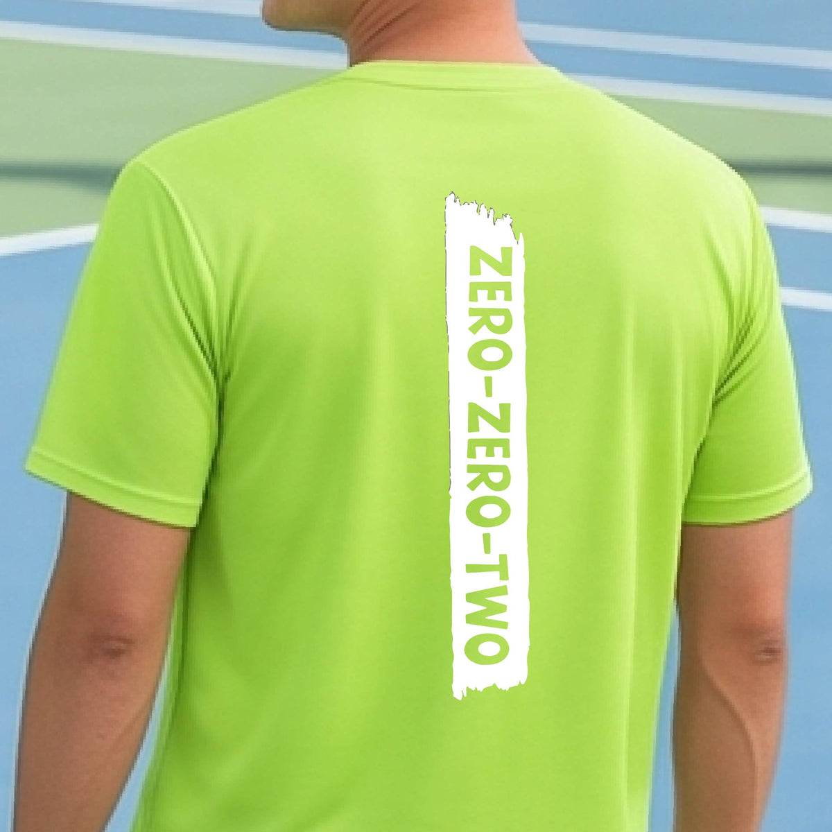 A close-up of a man on a pickleball wearing a lime green short-sleeve t-shirt, with a white graphic of artistic banner with the cutout words of ‘ZERO ZERO TWO’, on the back of the shirt. 