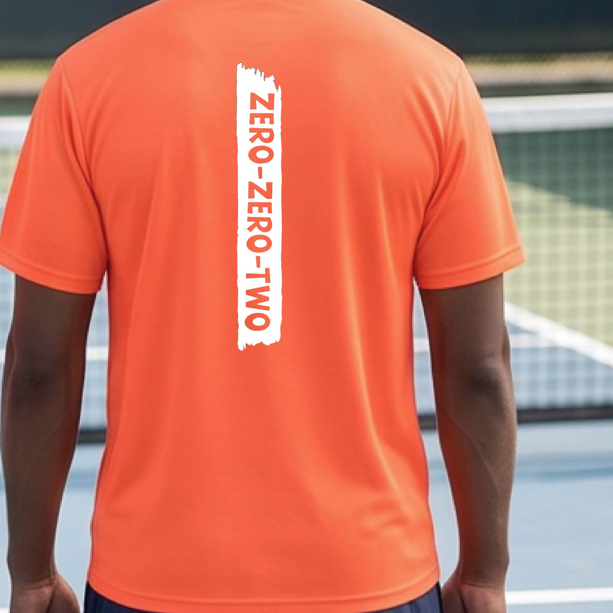 A close-up of a man on a pickleball wearing a orange short-sleeve t-shirt, with a white graphic of artistic banner with the cutout words of ‘ZERO ZERO TWO’, on the back of the shirt. 