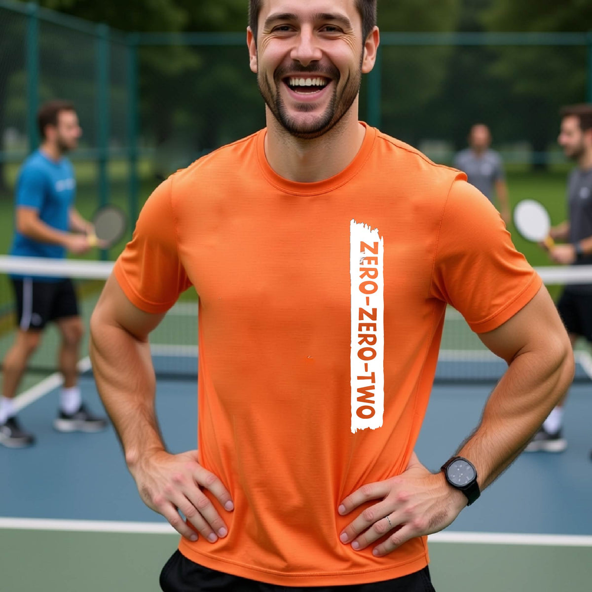 A close-up of a man on a pickleball wearing a orange short-sleeve t-shirt, with a white graphic of artistic banner with the cutout words of ‘ZERO ZERO TWO’, on the front of the shirt. 