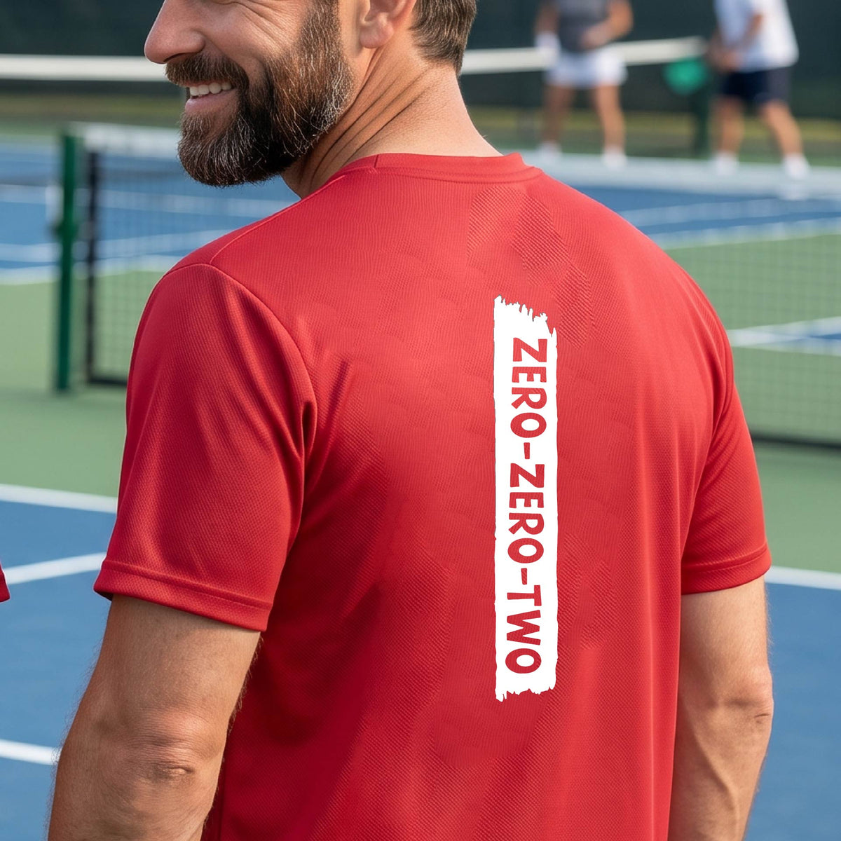 A close-up of a man on a pickleball wearing a red short-sleeve t-shirt, with a white graphic of artistic banner with the cutout words of ‘ZERO ZERO TWO’, on the back of the shirt. 