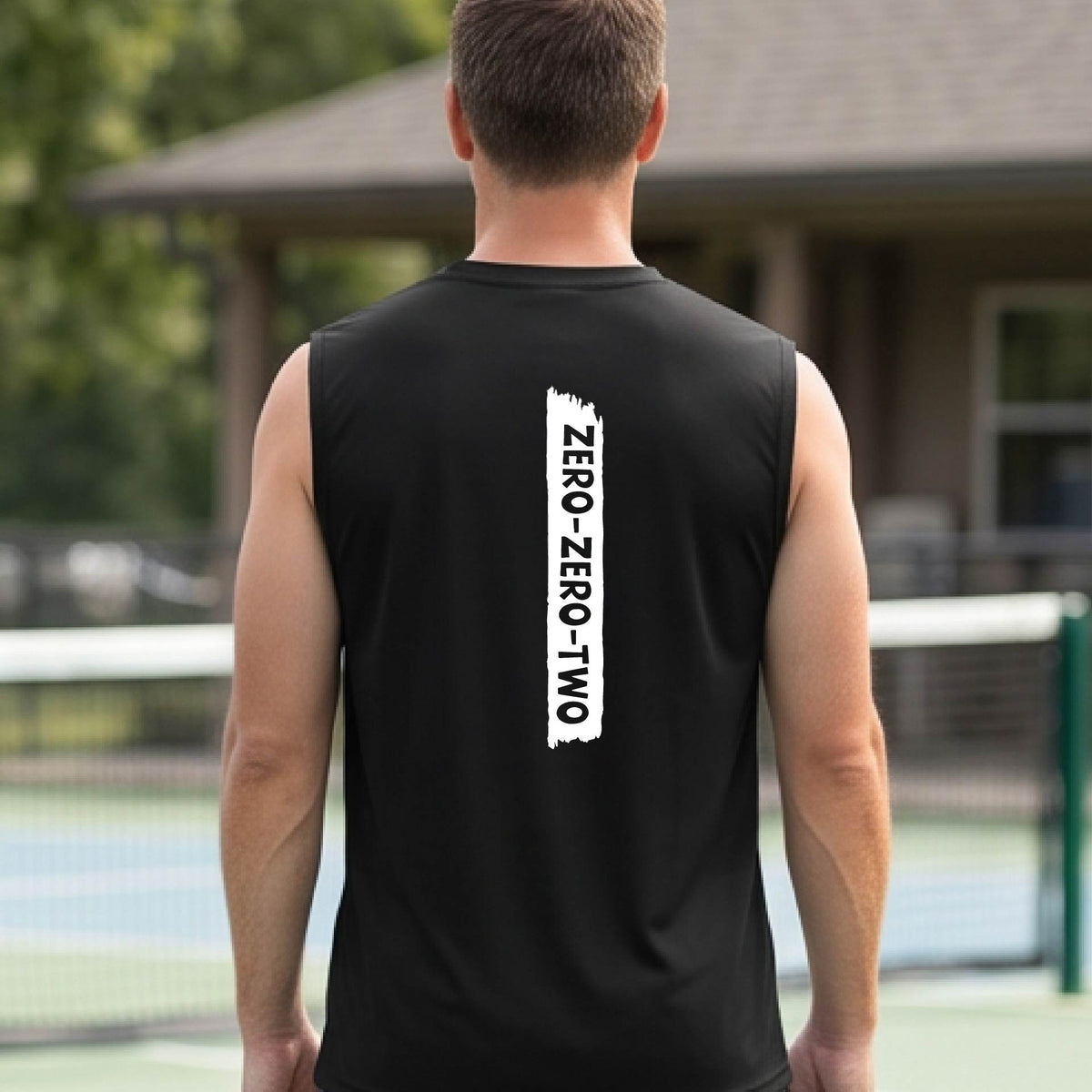 A close-up of a man on a pickleball wearing a black sleeveless t-shirt with a white graphic of artistic banner with the cutout words of ‘ZERO ZERO TWO’, on the back of the shirt. 