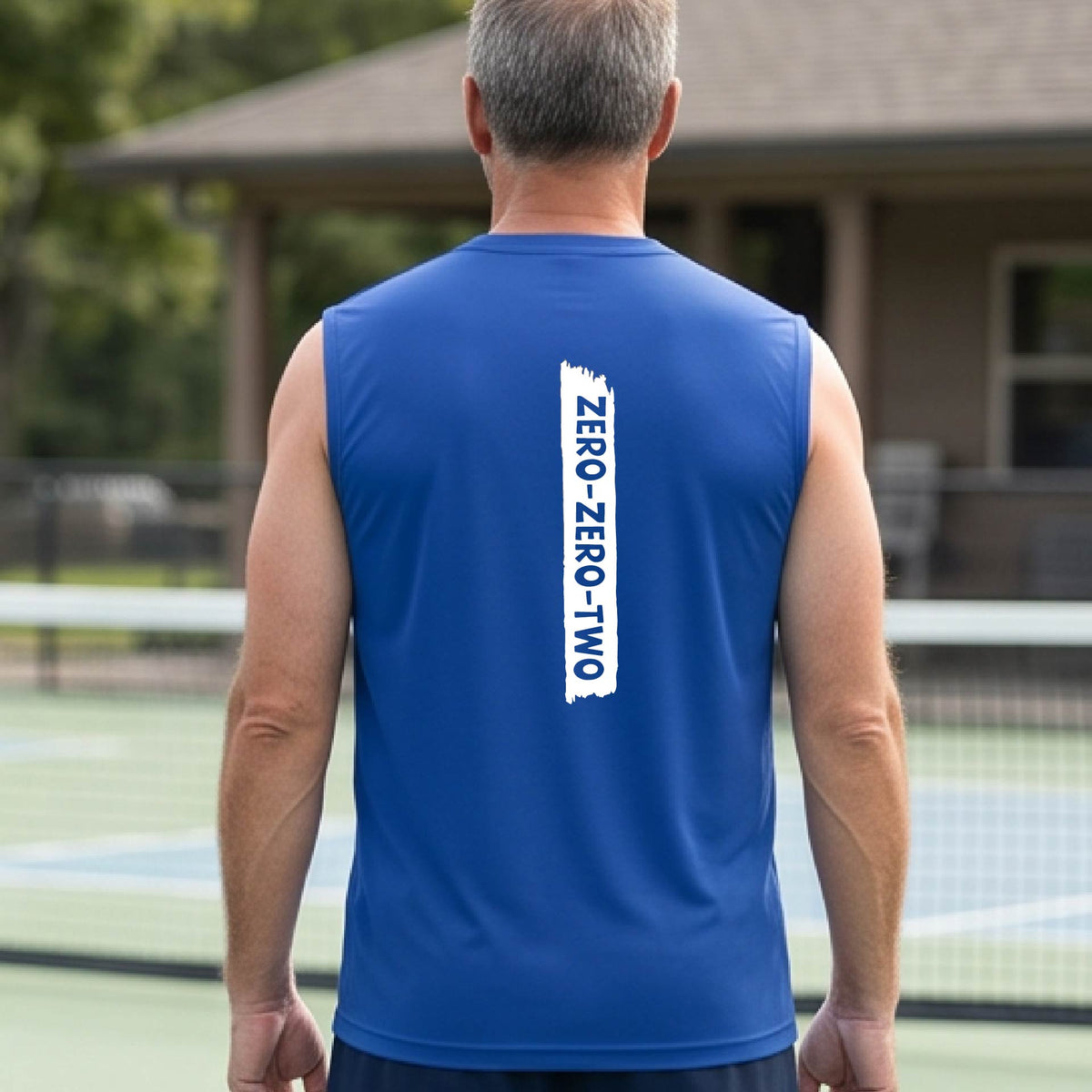 A close-up of a man on a pickleball wearing a royal bluesleeveless t-shirt with a white graphic of artistic banner with the cutout words of ‘ZERO ZERO TWO’, on the back of the shirt. 