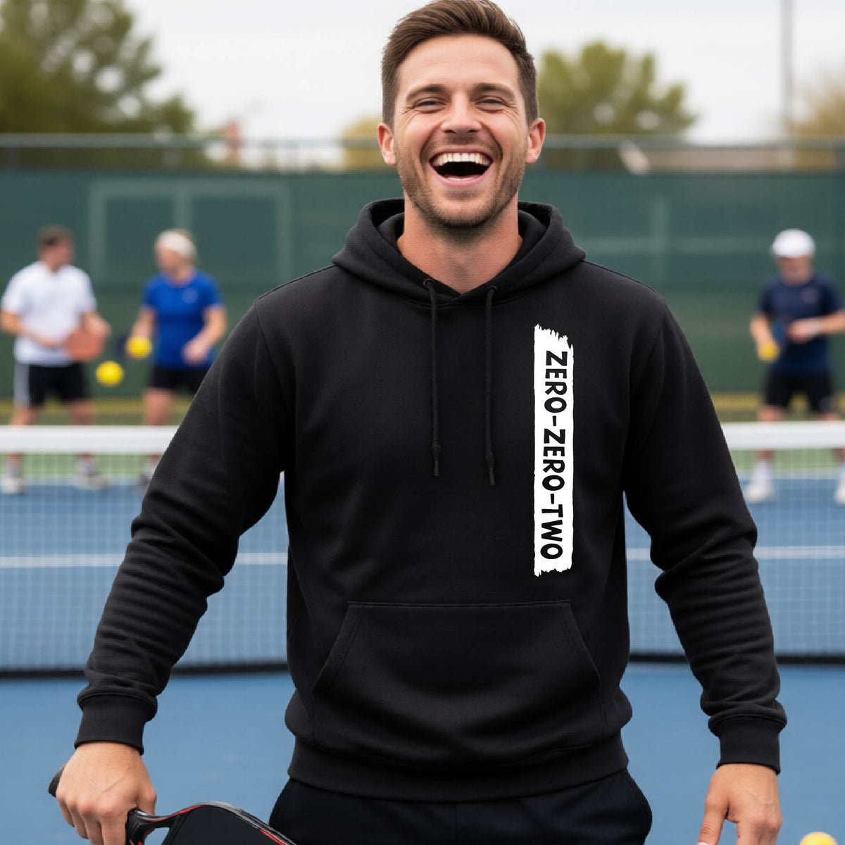 A close-up of a man on a pickleball court wearing a black hoodie pullover, with a white graphic of artistic banner with the cutout words of ‘ZERO ZERO TWO’, on the front of the hoodie. 