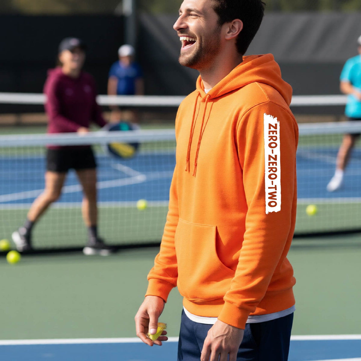 A close-up of a man on a pickleball court wearing an orange hoodie pullover, with a white graphic of artistic banner with the cutout words of ‘ZERO ZERO TWO’, on the sleeve of the hoodie. 