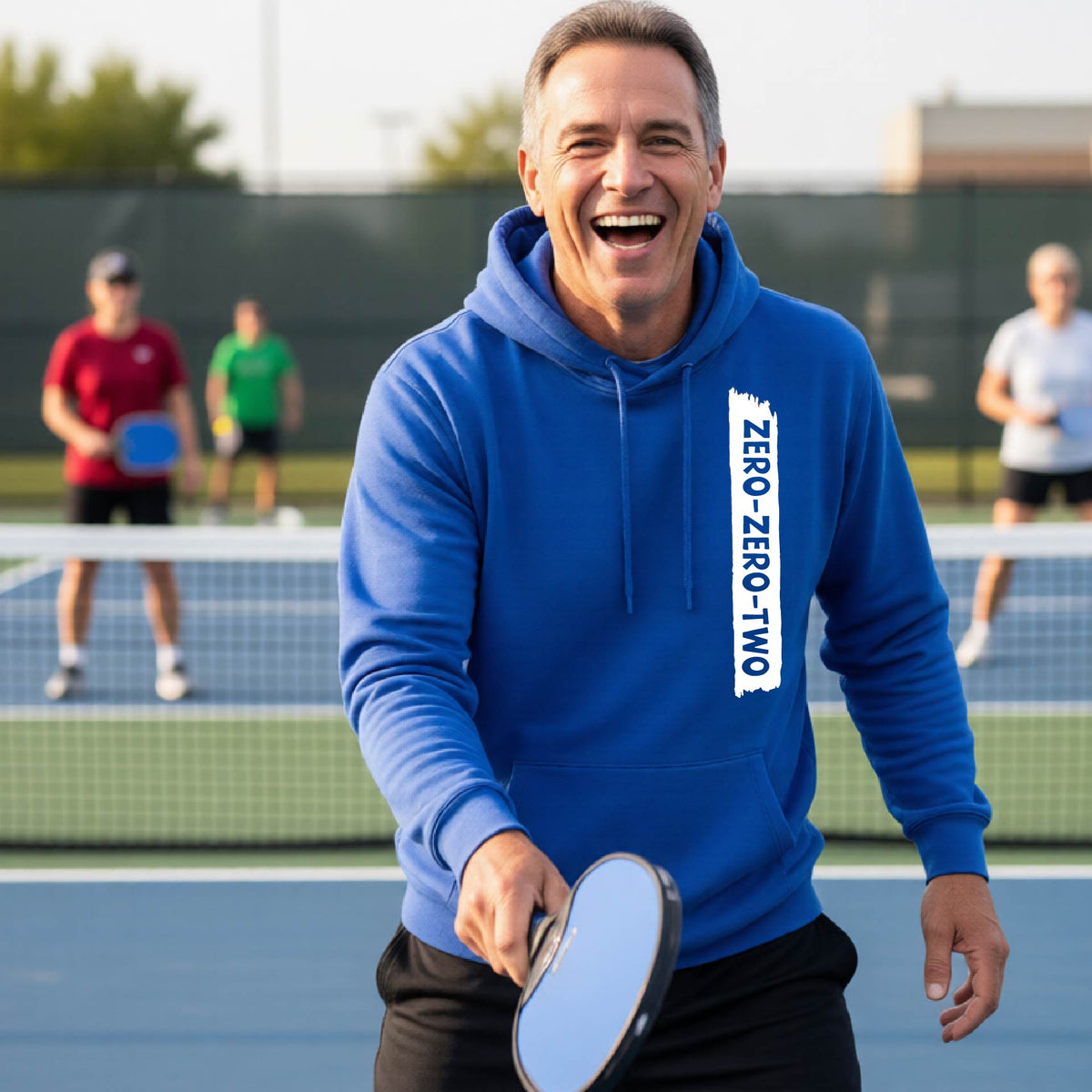 A close-up of a man on a pickleball court wearing a royal blue hoodie pullover, with a white graphic of artistic banner with the cutout words of ‘ZERO ZERO TWO’, on the front of the hoodie. 