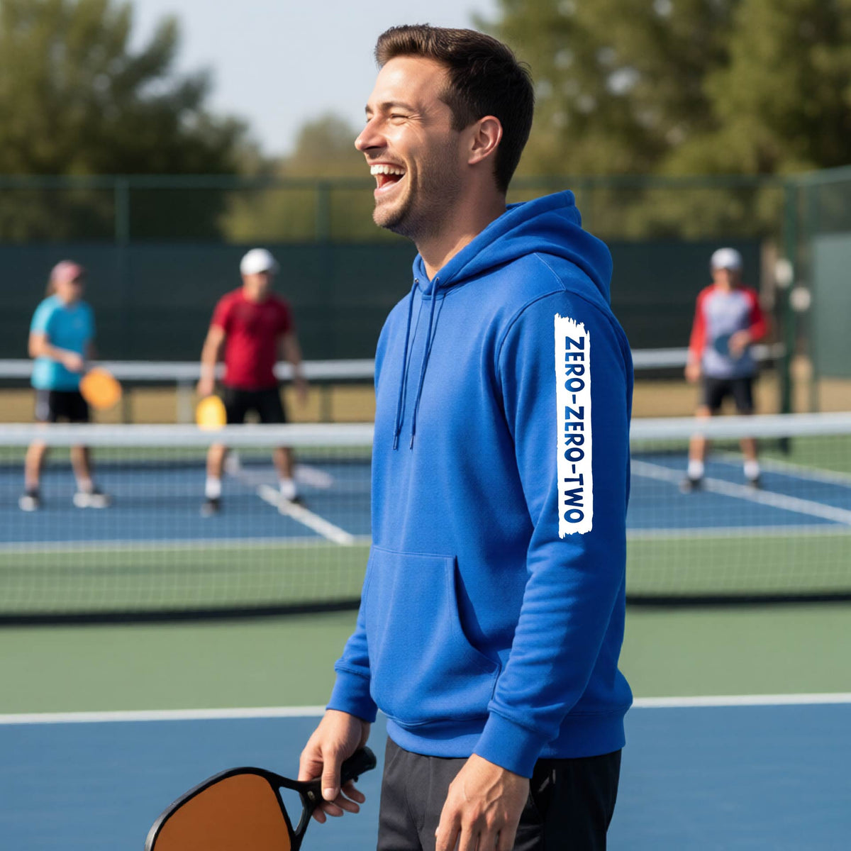 A close-up of a man on a pickleball court wearing a royal blue hoodie pullover, with a white graphic of artistic banner with the cutout words of ‘ZERO ZERO TWO’, on the sleeve of the hoodie. 