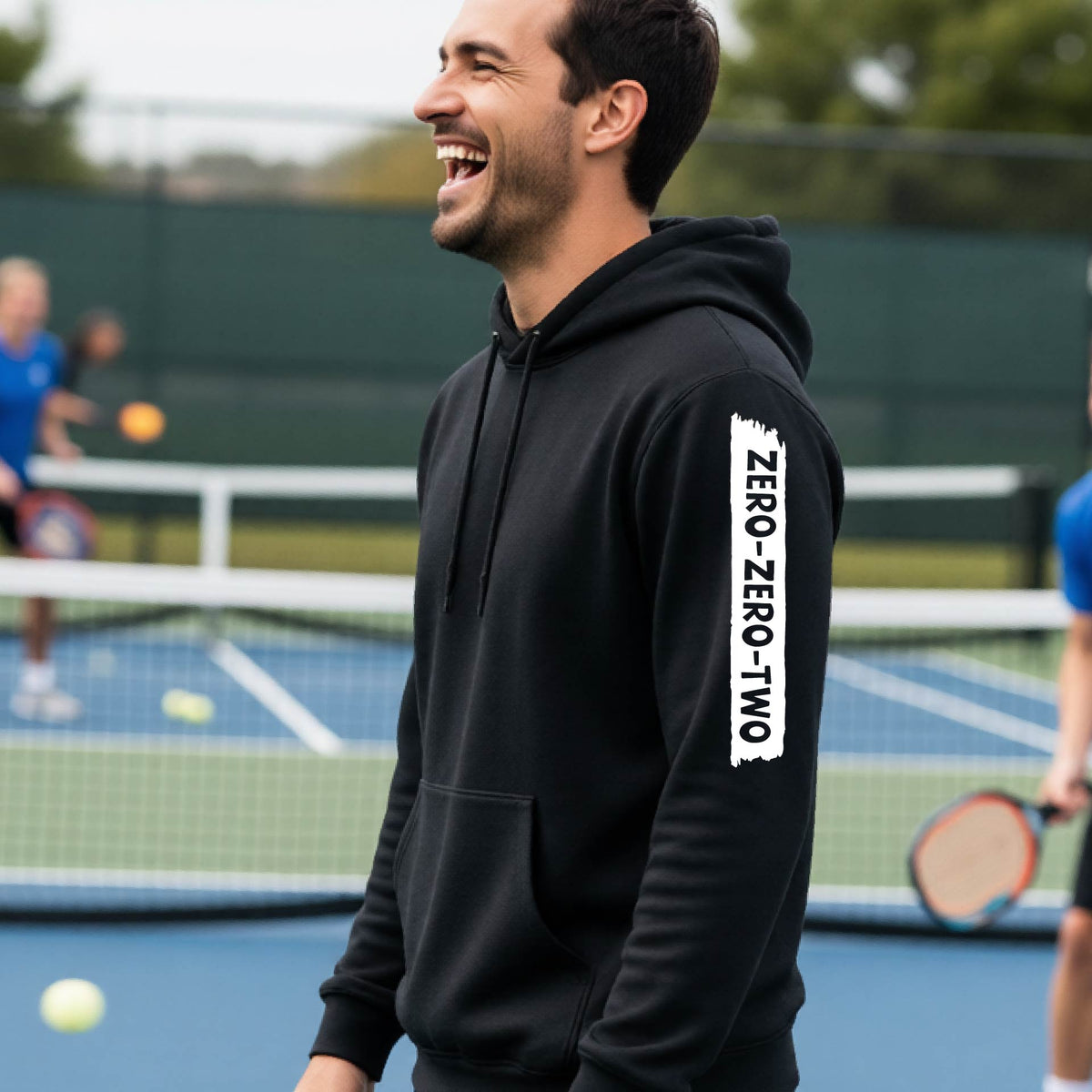 A close-up of a man on a pickleball court wearing a black hoodie pullover, with a white graphic of artistic banner with the cutout words of ‘ZERO ZERO TWO’, on the sleeve of the hoodie. 