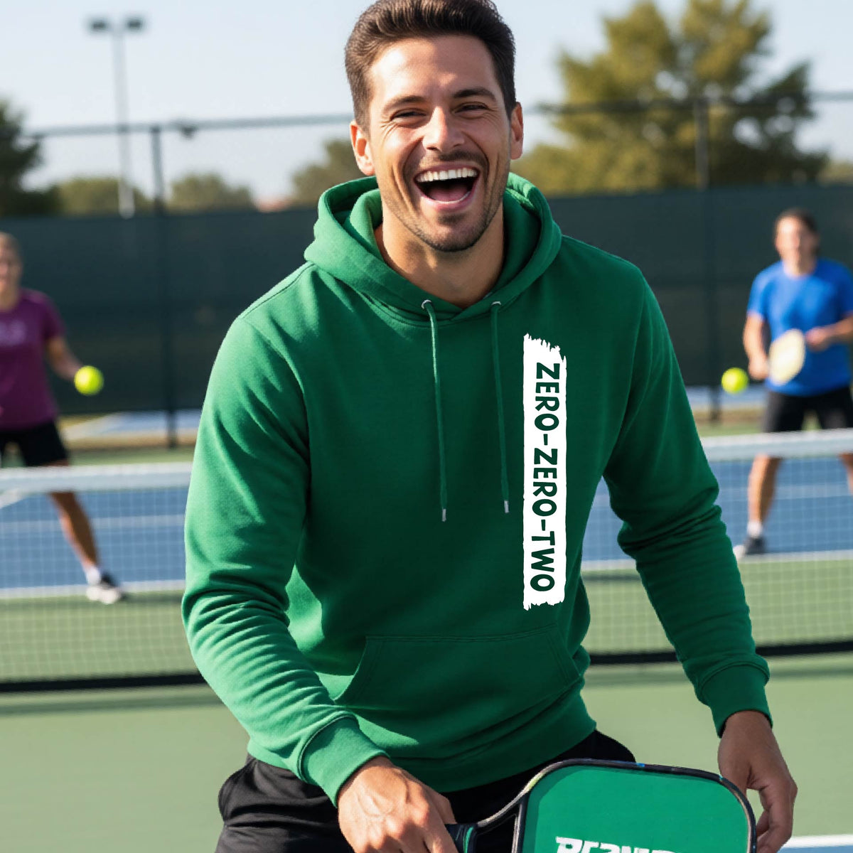 A close-up of a man on a pickleball court wearing a green hoodie pullover, with a white graphic of artistic banner with the cutout words of ‘ZERO ZERO TWO’, on the front of the hoodie. 