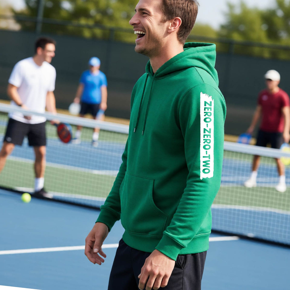 A close-up of a man on a pickleball court wearing a green hoodie pullover, with a white graphic of artistic banner with the cutout words of ‘ZERO ZERO TWO’, on the sleeve of the hoodie. 