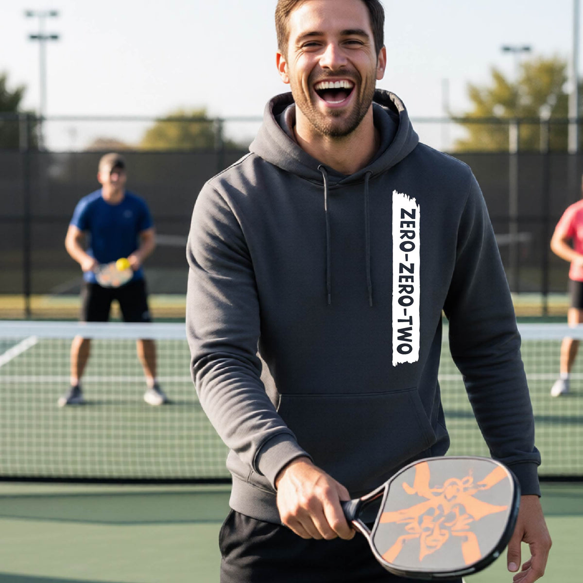 A close-up of a man on a pickleball court wearing a gray hoodie pullover, with a white graphic of artistic banner with the cutout words of ‘ZERO ZERO TWO’, on the front of the hoodie. 