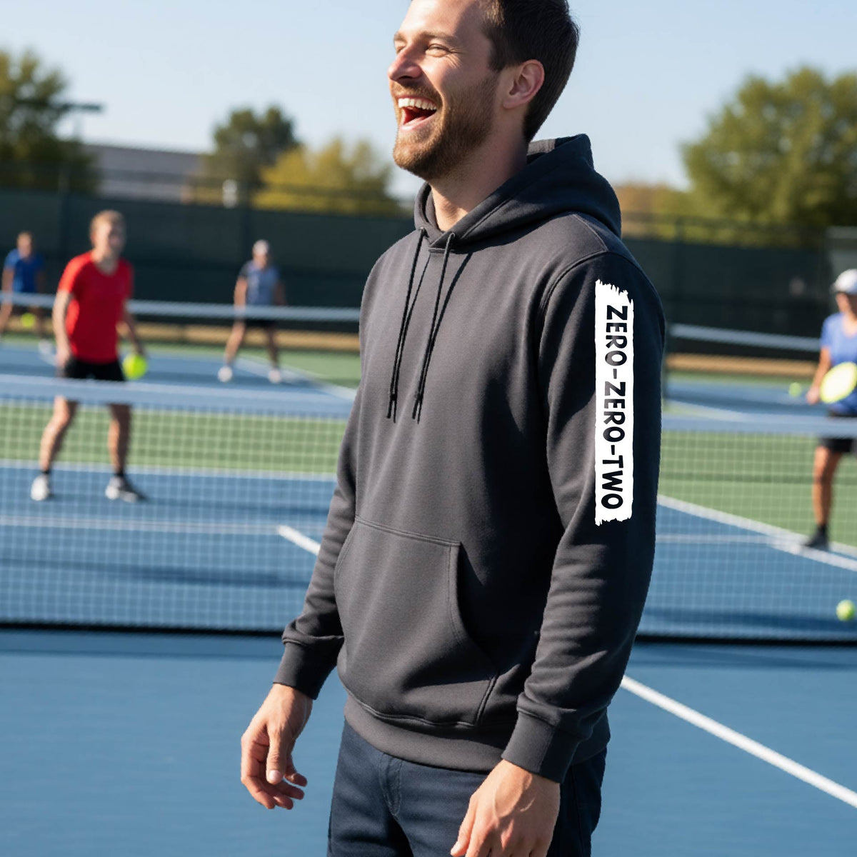 A close-up of a man on a pickleball court wearing a gray hoodie pullover, with a white graphic of artistic banner with the cutout words of ‘ZERO ZERO TWO’, on the sleeve of the hoodie. 