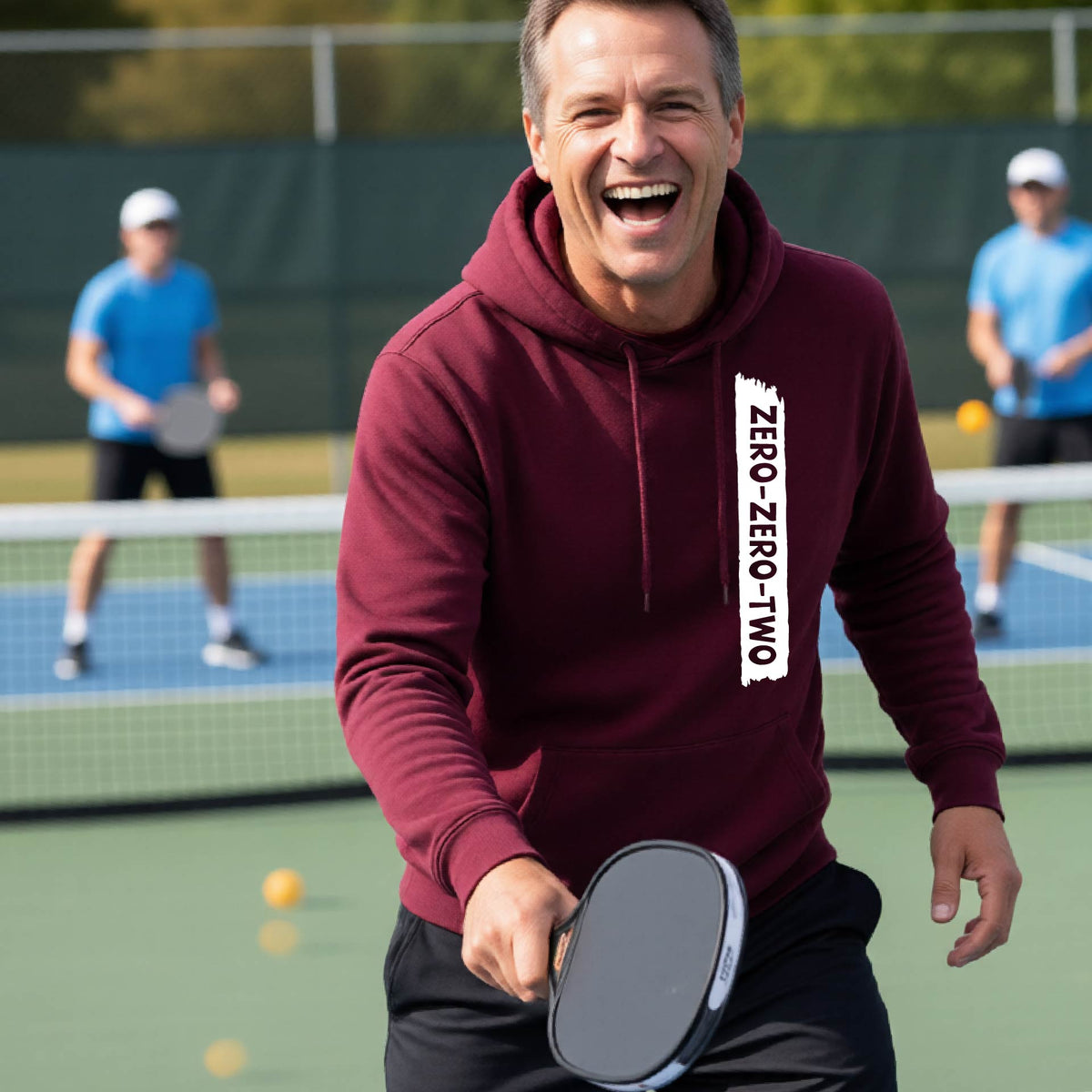 A close-up of a man on a pickleball court wearing a maroon hoodie pullover, with a white graphic of artistic banner with the cutout words of ‘ZERO ZERO TWO’, on the front of the hoodie. 