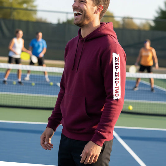 A close-up of a man on a pickleball court wearing a maroon hoodie pullover, with a white graphic of artistic banner with the cutout words of ‘ZERO ZERO TWO’, on the sleeve of the hoodie. 