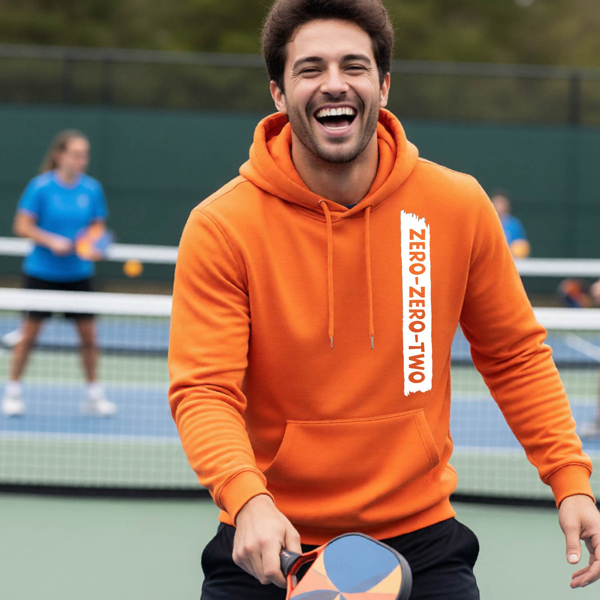 A close-up of a man on a pickleball court wearing an orange hoodie pullover, with a white graphic of artistic banner with the cutout words of ‘ZERO ZERO TWO’, on the front of the hoodie. 