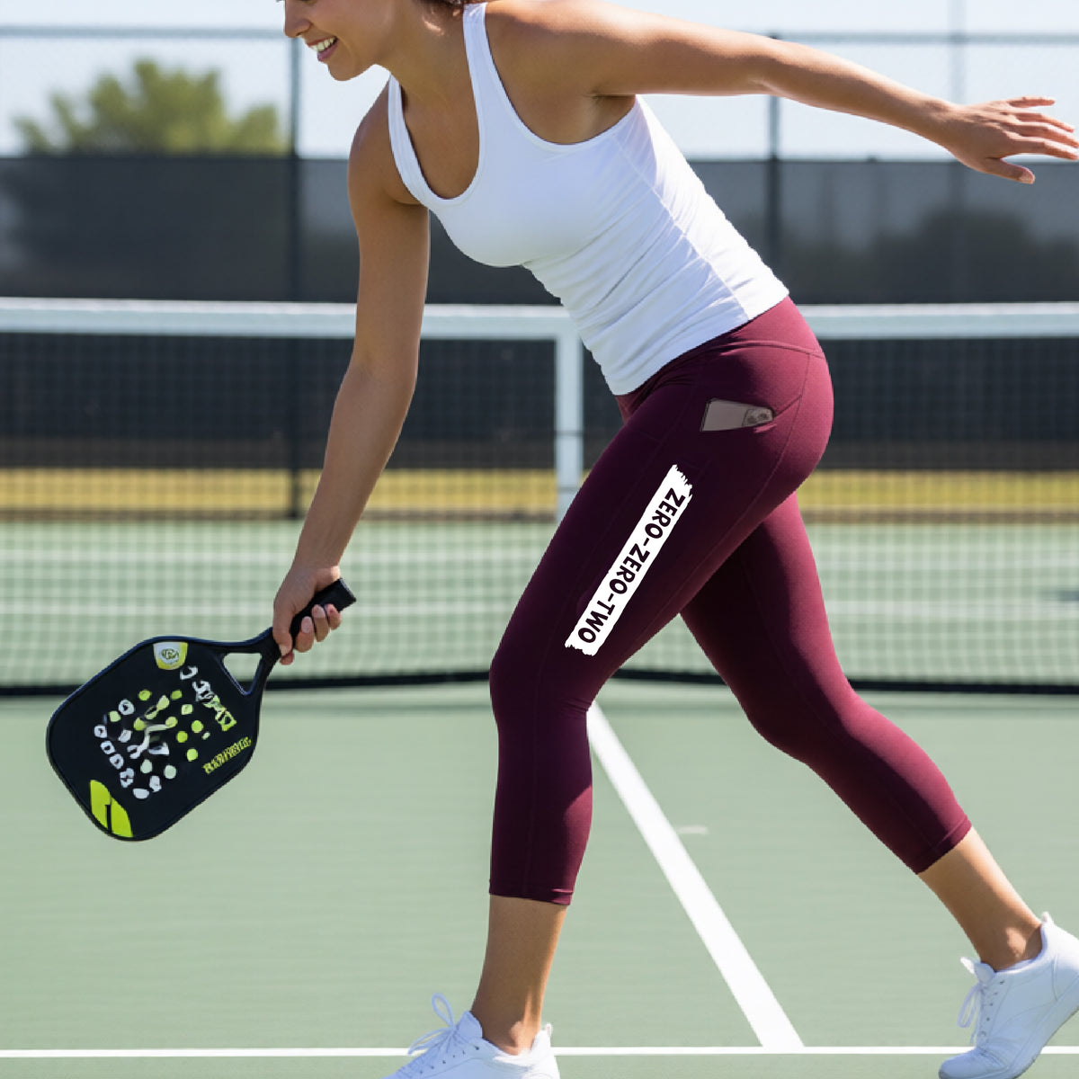 A woman on a pickleball court wearing maroon colored capris and a white graphic down the leg of white graphic of artistic banner with the cutout words of ‘ZERO ZERO TWO’ , on the capris.