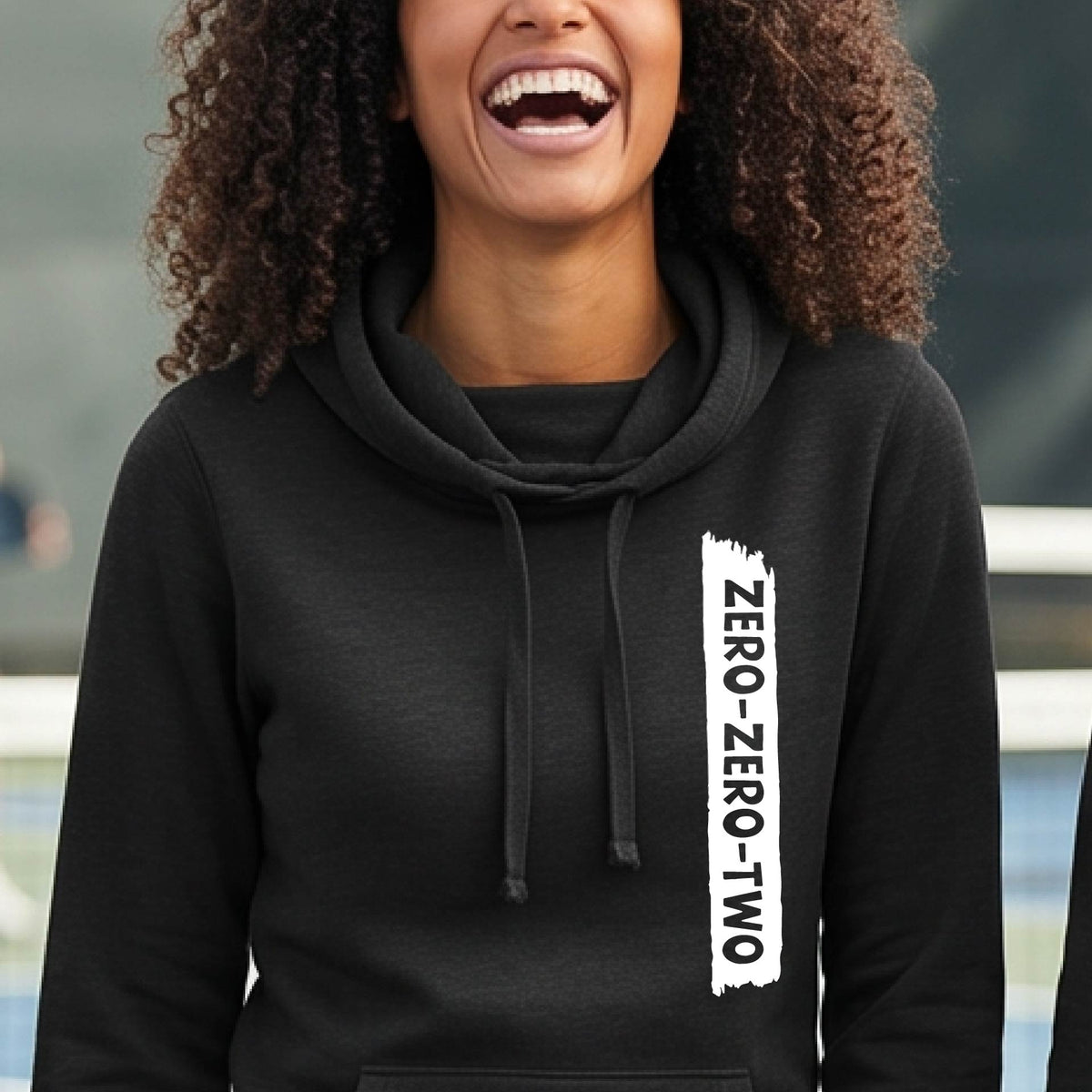 A close-up of a woman on a pickleball court wearing a black cowl-neck hoodie pullover, with a white graphic of artistic banner with the cutout words of ‘ZERO ZERO TWO’ , on the front of the hoodie. 