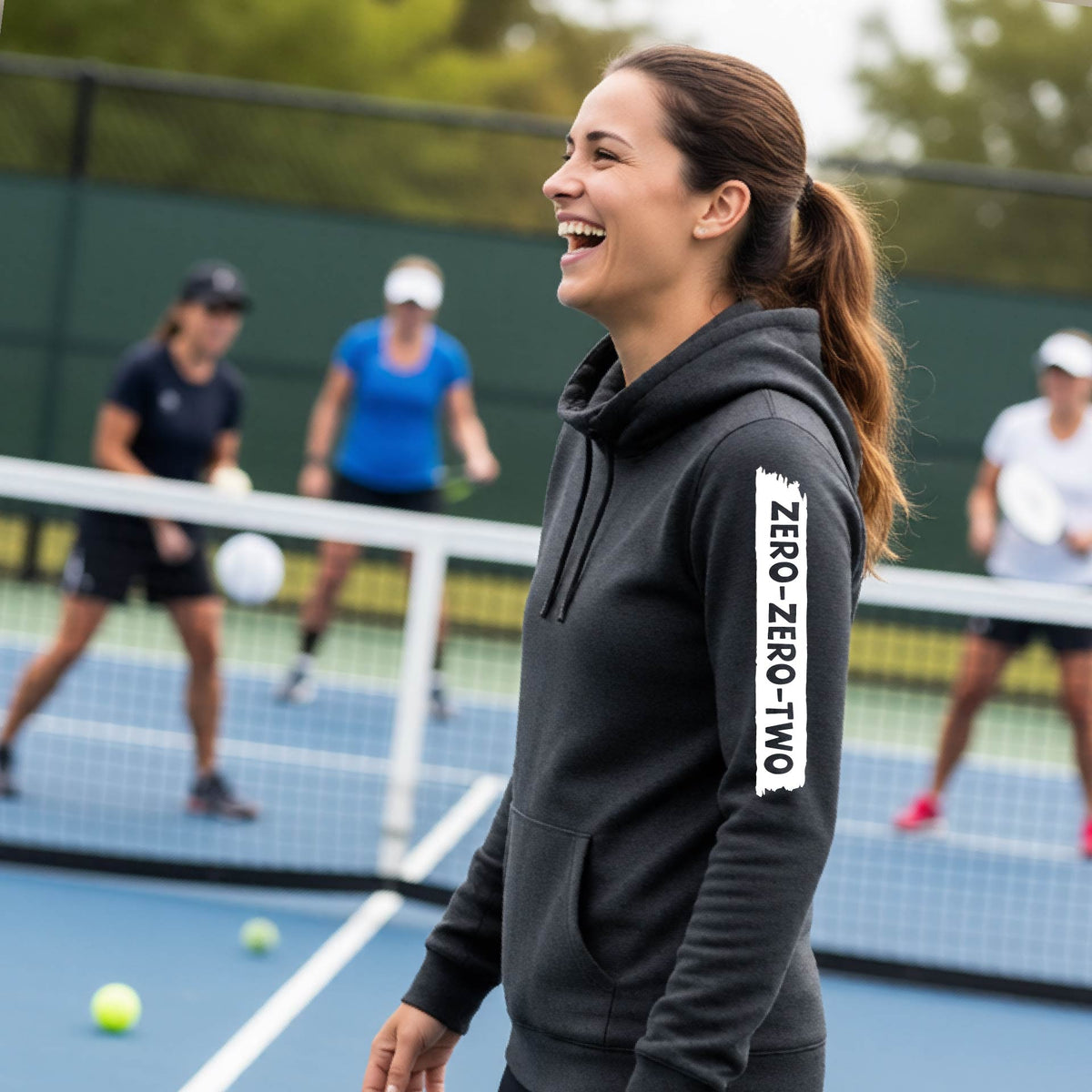 A close-up of a woman on a pickleball court wearing a black cowl-neck hoodie pullover, with a white graphic of artistic banner with the cutout words of ‘ZERO ZERO TWO’ , on the sleeve of the hoodie. 