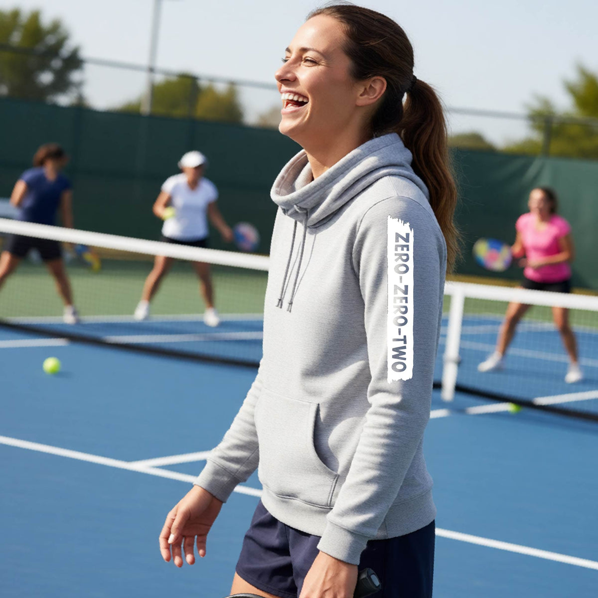 A close-up of a woman on a pickleball court wearing a light gray  cowl-neck hoodie pullover, with a white graphic of artistic banner with the cutout words of ‘ZERO ZERO TWO’ , on the sleeve of the hoodie. 