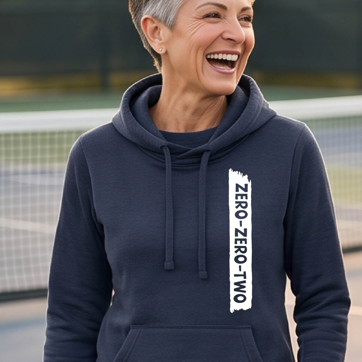 A close-up of a woman on a pickleball court wearing a navy blue  cowl-neck hoodie pullover, with a white graphic of artistic banner with the cutout words of ‘ZERO ZERO TWO’ , on the front of the hoodie. 