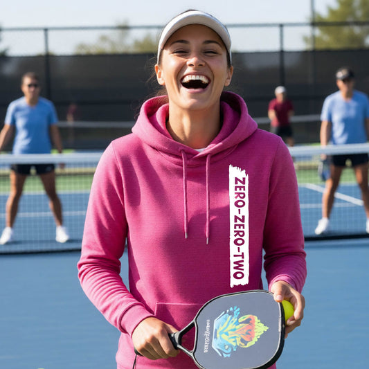 A close-up of a woman on a pickleball court wearing a pink cowl-neck hoodie pullover, with a white graphic of artistic banner with the cutout words of ‘ZERO ZERO TWO’ , on the the front of the hoodie. 