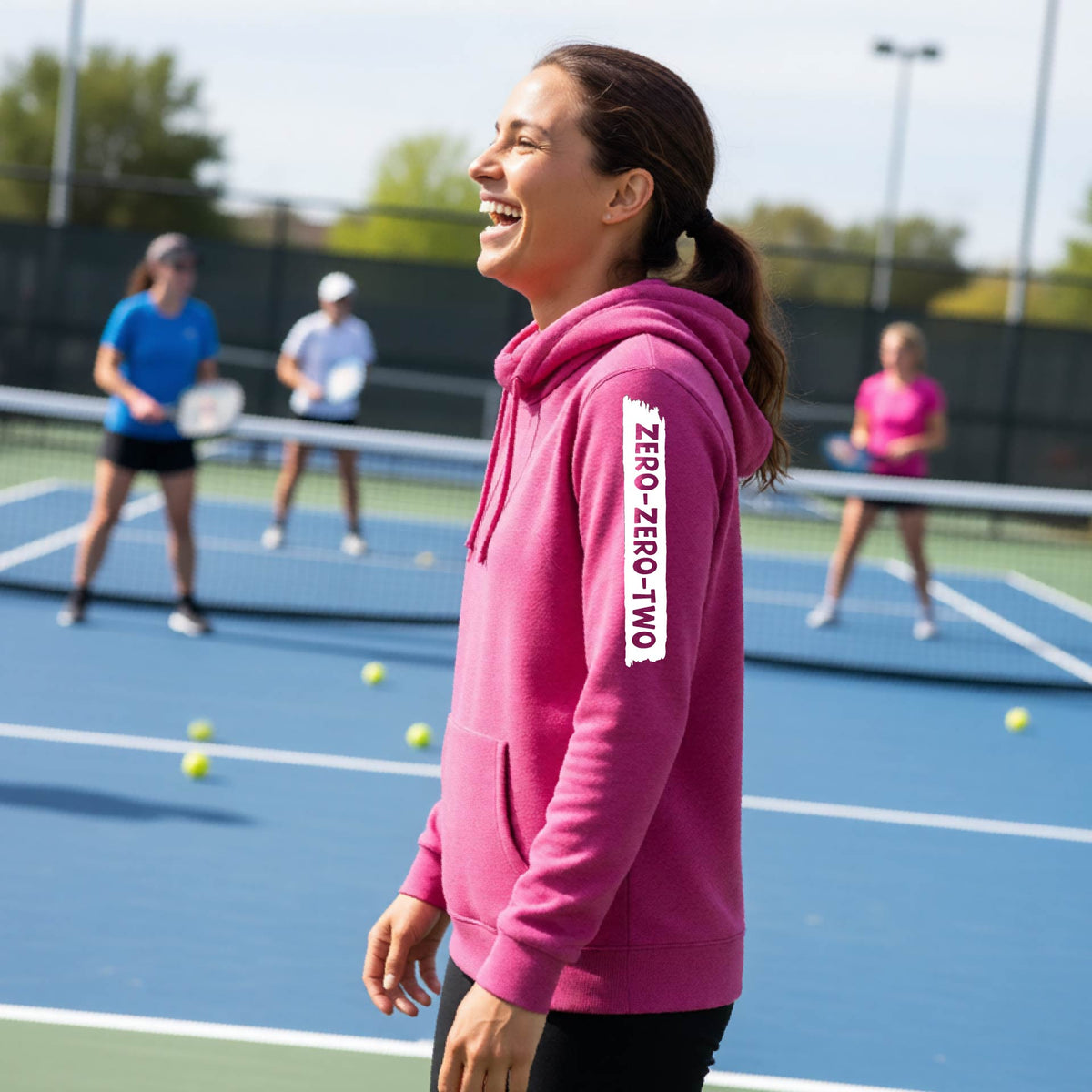 A close-up of a woman on a pickleball court wearing a pink cowl-neck hoodie pullover, with a white graphic of artistic banner with the cutout words of ‘ZERO ZERO TWO’ , on the sleeve of the hoodie. 
