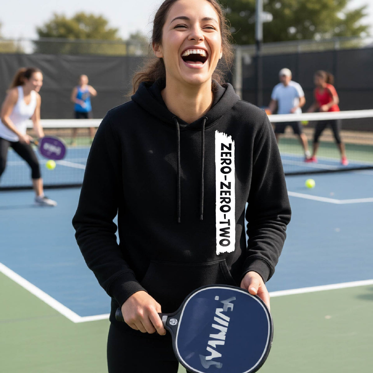 A close-up of a woman on a pickleball court wearing a black fitted hoodie pullover, with a white graphic of artistic banner with the cutout words of ‘ZERO ZERO TWO’ , on the front of the  hoodie. 